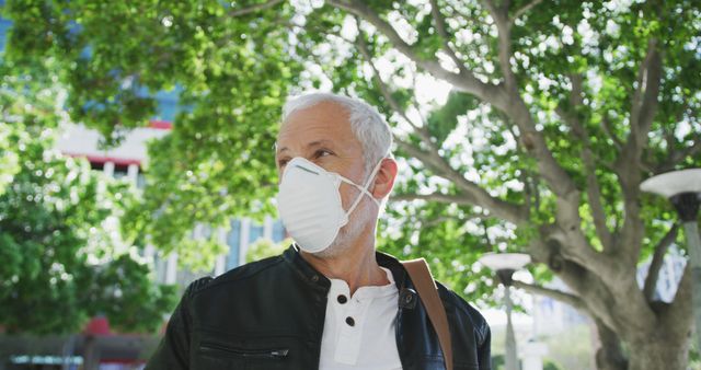 Senior man wearing a face mask for protection while outdoors, shown during daylight under a tree. Ideal for health safety campaigns, pandemic-related visuals, promoting outdoor activities with precautions, and lifestyle content featuring older adults.