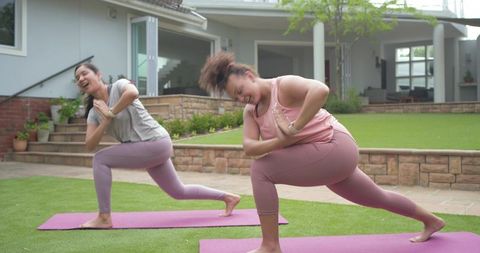 Diverse women practicing yoga on backyard mats in athletic wear, laughing during low lunge twist