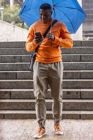 African american man holding blue umbrella checking smartphone on rainy city steps