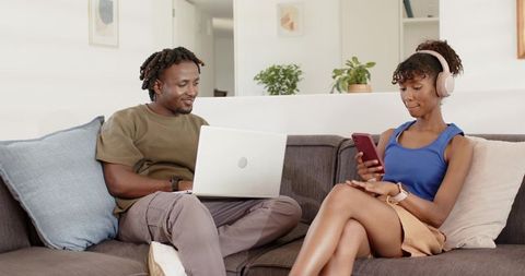Young couple relaxing on couch using laptop and smartphone with wireless headphones