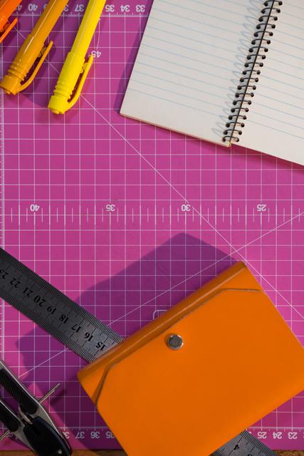 This image shows various school supplies arranged on a pink cutting mat. Items include a spiral notebook, yellow pens, a metal ruler, an orange pouch, and a hole puncher. Ideal for use in educational materials, back-to-school promotions, organizational tips, and creative workspace inspiration.