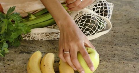 African american woman reaching for green apple with reusable mesh bag on granite countertop