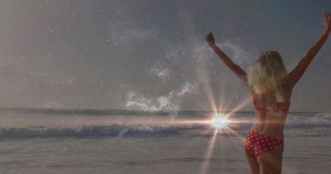 Woman raising arms facing ocean at sunset wearing red polka-dot bikini with sun flare