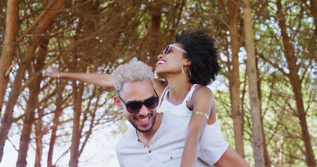 A young African American couple enjoys a playful moment outdoors, with the woman piggybacking on the man, both smiling broadly. Their joyful expressions and casual summer attire suggest a carefree, romantic day spent together.