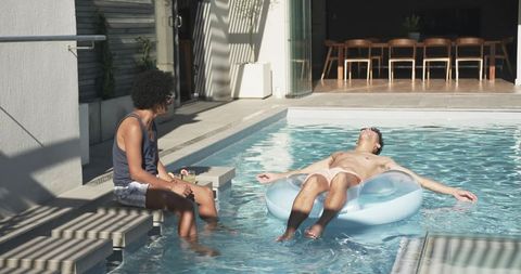 Two men relaxing by pool floating on inflatable ring and sipping drinks backyard summer