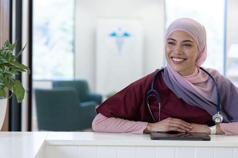 Smiling nurse wearing headscarf at clinic reception desk using tablet and stethoscope