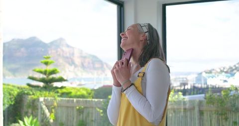 Mature woman smiling and holding smartphone by sunlit coastal window with harbor view