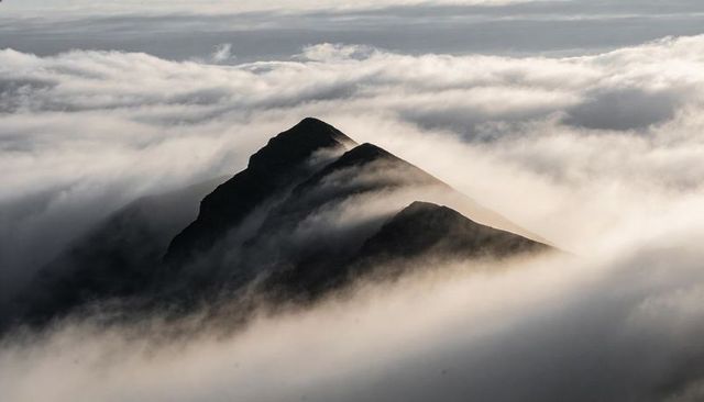 Sunlit jagged mountain ridge rising above sea of clouds at high elevation, dramatic fog