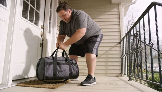 Middle-aged man lifting heavy gray duffel on covered porch while preparing for travel