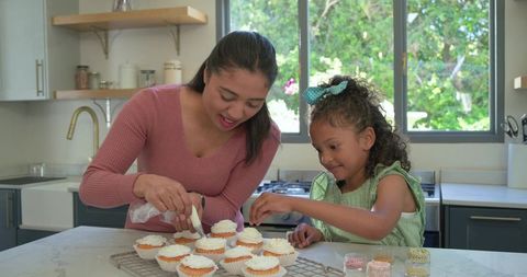 Mother and daughter decorating cupcakes together on sunlit modern kitchen island
