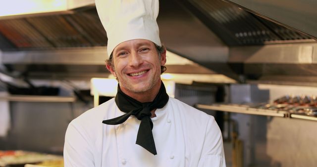 Friendly chef wearing a traditional white uniform with a hat and black necktie, standing in a professional kitchen and smiling warmly. This can be used for advertising restaurants, culinary schools, cooking classes, and food blogs.