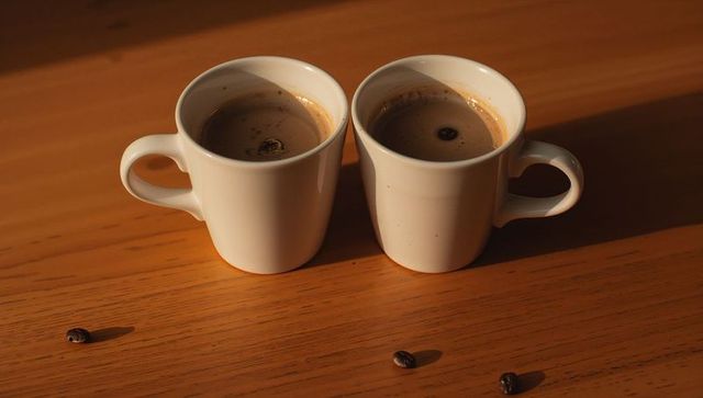Two white espresso cups resting on sunlit wooden table with crema and coffee beans