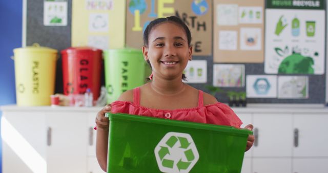 Smiling girl holding a green recycling bin in a classroom centered on environmental education. Posters and bins for different materials like plastic, paper, and glass are visible in the background. Ideal for use in educational content about recycling, sustainability programs, eco-friendly initiatives, or promoting environmental consciousness among children.