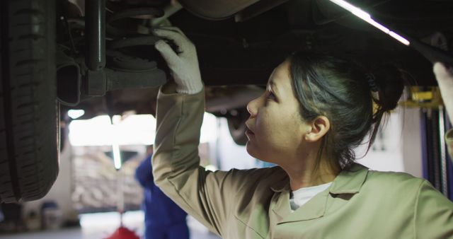 A female auto mechanic is inspecting the undercarriage of a vehicle in a well-lit garage. She is wearing protective gloves and mechanical uniform, focusing intently on her work. This image illustrates the role and dedication of women in the automotive service industry. Useful for promoting gender diversity in trade professions, automotive service advertisements, educational materials on car maintenance, and career guidance resources.