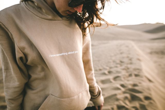 Young woman with brown hair wearing beige hoodie with text 'I know something about you' and sunglasses, standing in desert sand dunes. Hair is blowing in the wind. Could be used for advertisements, social media content, blogs, or articles about fashion, travel, or desert environments.