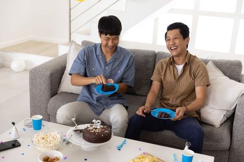 Asian male relatives sharing chocolate cake on sofa laughing during home celebration