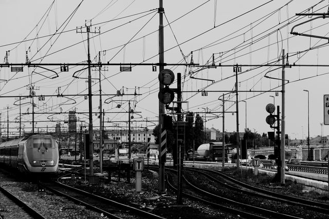Monochrome image depicting urban railway tracks with complex overhead electrical wires, suggesting a busy city environment. Can be used in urban studies, transportation, and infrastructure presentations or publications.
