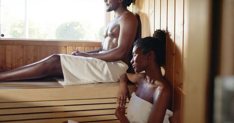 African american couple relaxing in sunlit wooden sauna wearing towels
