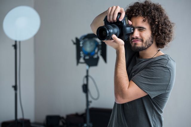 Male photographer with curly hair is using a digital camera in a photography studio. He looks happy and focused on taking pictures. This image is suitable for websites, articles, or advertisements related to photography, professional photographers, studio equipment, or hobbyists taking pictures.