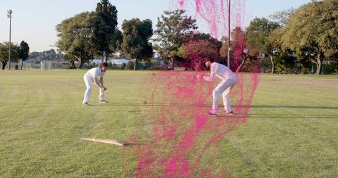 Two cricketers fielding on lakeside grass wearing white kit with pink paint splash