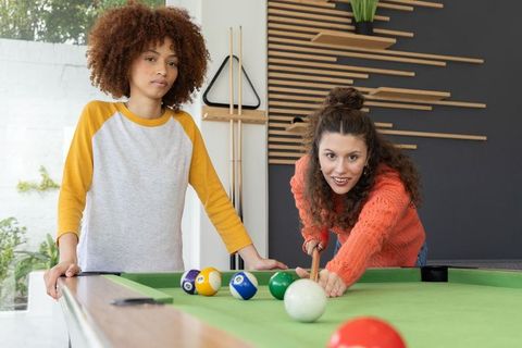 Teen girl and woman playing billiards, woman lining up cue shot while friend watching