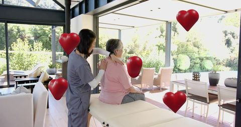 Home caregiver giving gentle shoulder therapy to senior woman on treatment table with heart balloons