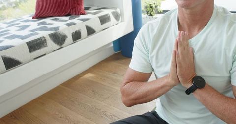 Mature man kneeling and meditating by window bench on hardwood floor with smartwatch