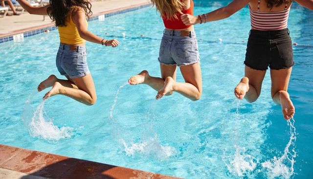 Three friends jumping and splashing into sunny outdoor pool wearing tanks and denim shorts