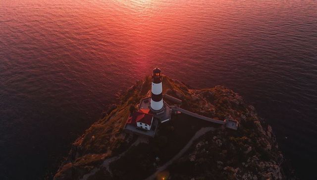 Sunset striped lighthouse on rocky headland with red-roof keeper house, aerial coastline