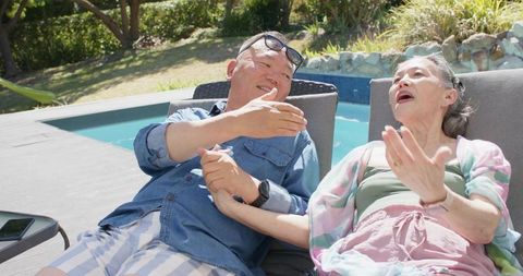 Asian senior couple relaxing and holding hands on poolside loungers enjoying sunny backyard