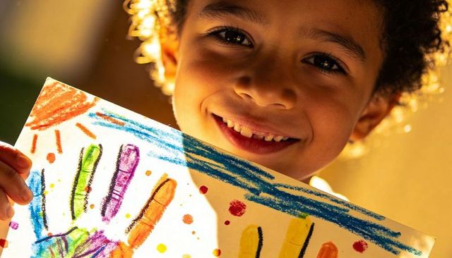 Smiling curly-haired child holding colorful handprint art in golden sunlight