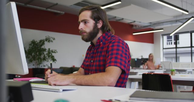 Man at contemporary workspace focusing on a computer while taking notes. Ideal for themes on work environments, corporate culture, productivity, modern offices, and young professionals.