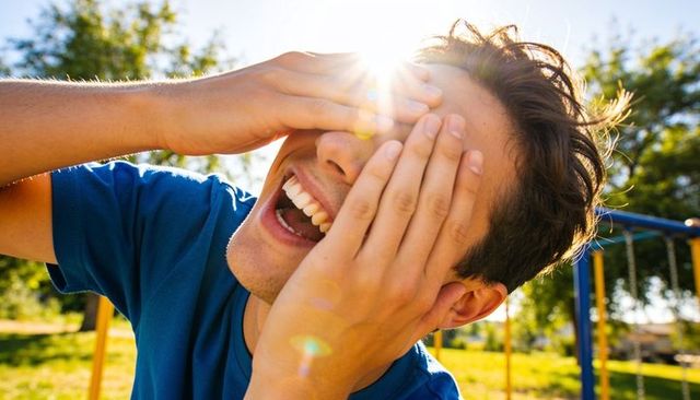 Laughing young man covering eyes with hands while sun flaring over playground