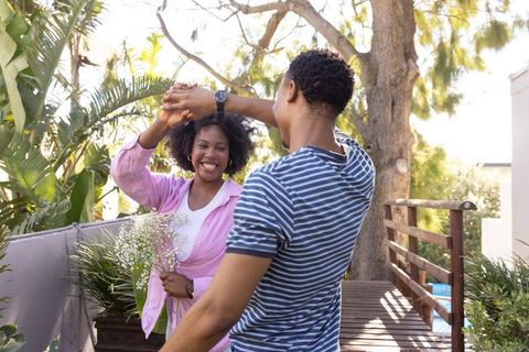 Sunlit african american couple dancing on terrace while man twirling woman with bouquet