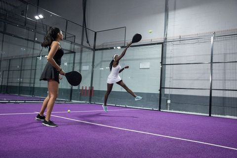 Diverse female players leaping and smashing padel on purple indoor court for team play