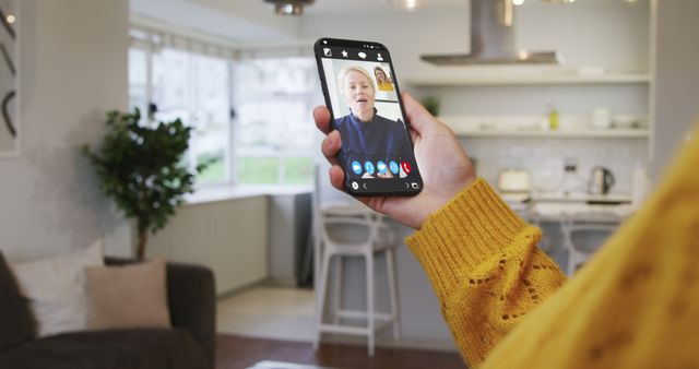 Person in yellow sweater using smartphone for video call in modern kitchen. Modern design with natural light. Perfect for online communication, staying connected with family, and remote working concepts.