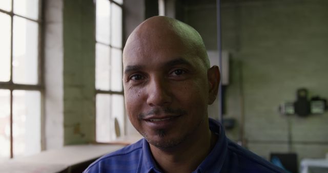 Bald man with a friendly smile standing indoors in an industrial environment with natural light coming through large windows. Suitable for use in workplace diversity campaigns, industrial work promotions, or human interest pieces focusing on factory workers.