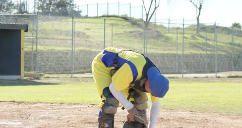 Baseball catcher adjusting protective gear at home plate wearing yellow jersey and blue helmet