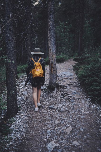 Ideal for illustrating themes of adventure, self-discovery, and nature exploration, this photo shows a woman hiking alone in a forest. The yellow backpack adds a vibrant contrast to the greens and browns of the surrounding woods, while her casual attire and hat emphasize casual outdoor activity. Perfect for nature blogs, travel articles, and advertisements promoting outdoor gear and adventure tourism.