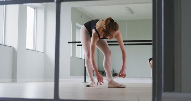 Ballet dancer stretching in light-filled dance studio. Ideal for illustrating themes of dance, preparation, flexibility, and dedicated practice. Useful for promoting dance classes, fitness programs, or lifestyle blogs focused on professional training and health.