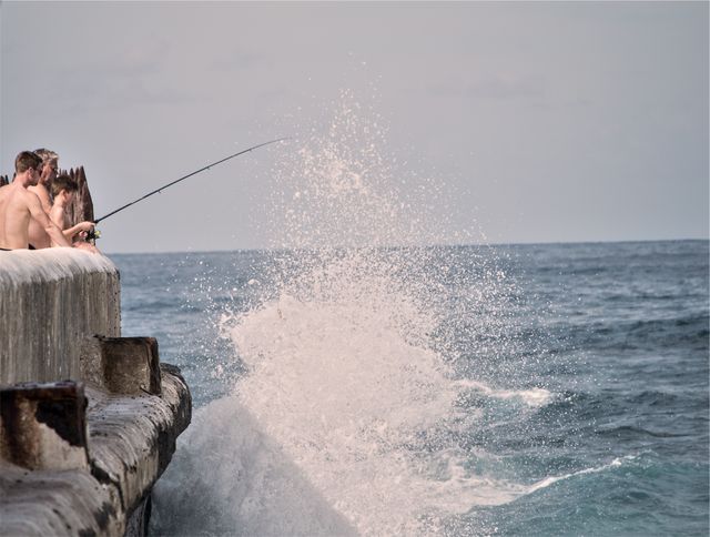 Men fishing on a seaside pier during daytime facing rough waves crashing against the structure. Perfect for content related to recreational fishing, outdoor adventures, summer activities, coastal experiences, and hobbies. Ideal for use in travel blogs, articles on leisure activities, fishing trip promotions, and lifestyle magazines.