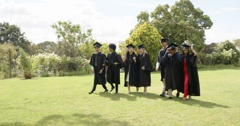 Diverse graduates walking on campus lawn in caps and gowns celebrating achievement