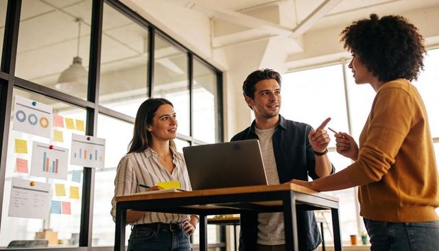Three colleagues collaborating around laptop at standing table in modern glass office