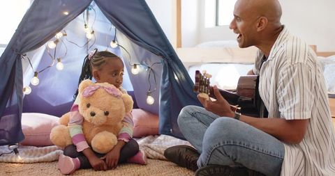 Mature father playing guitar with african american daughter hugging teddy in cozy bedroom tent