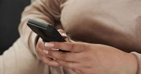 Mid-adult woman holding smartphone and typing with French manicure on beige sofa closeup home lifest