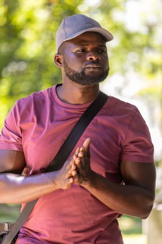 Urban man standing in sunlit park wearing gray cap, crossbody bag, pressing hands