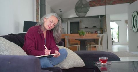 Senior woman writing journal on cozy contemporary sofa wearing cardigan in open-plan home