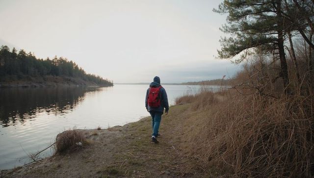Solitary hiker walking lakeside trail at dusk with red backpack