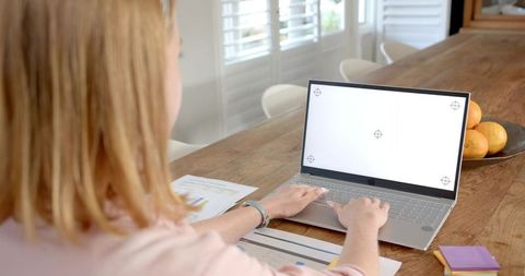 Woman working on laptop at wooden table reviewing financial charts in bright home workspace