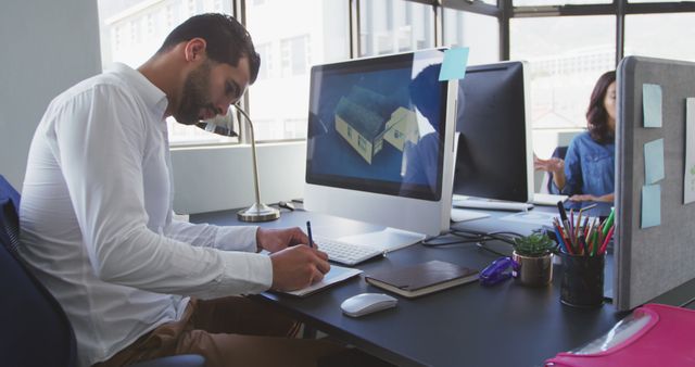 Young male architect working diligently on a design project at a modern, bright office environment. He is writing down notes while referring to a computer screen displaying architectural plans. In the background, a colleague focuses on her work at a nearby desk. Ideal for use in topics related to architecture, design professionals, modern workspaces, creative industries, and team collaboration.